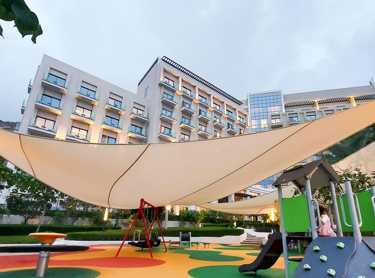 Ein Spielplatz mit Schaukeln und Rutschen, einem überdachten Sitzbereich und einem Gebäude im Hintergrund.
