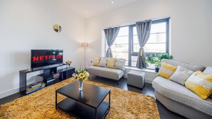 Ein modernes Wohnzimmer mit Fernseher, einer Sofagarnitur, einem Couchtisch und einem großen Fenster mit Vorhängen.