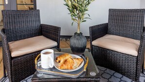 Zwei Korbstühle mit beige farbenen Kissen, ein Glastisch mit einer Auflage und einem Croissant sowie einer Tasse, und ein Blumentopf.