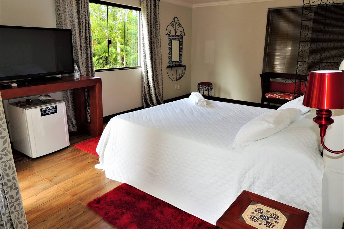 A hotel room with a bed, a TV, a small refrigerator, a red rug, and a window with curtains.