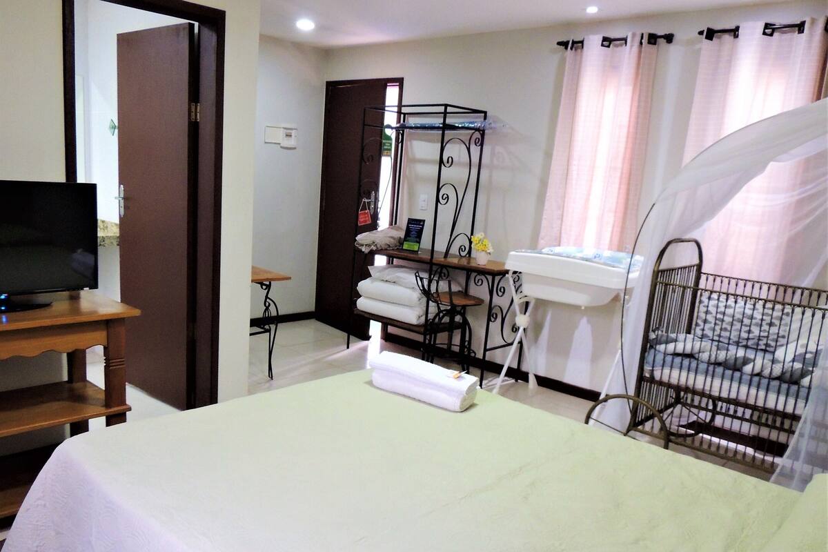 A hotel room with a bed, a television on a wooden table, a small table with a chair, a shelf with towels, a white sink, and a crib with a canopy.