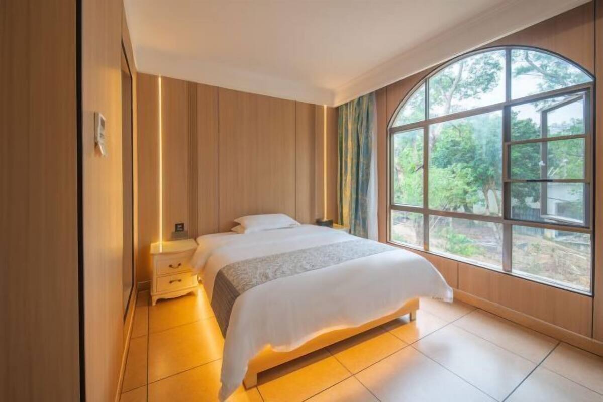 A bedroom with a large window, a bed, a bedside table, and a nightstand.