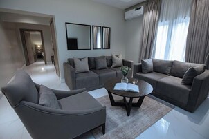 Uma sala de estar moderna com um sofá cinzento, uma mesa de centro com um livro e um vaso, e um espelho grande na parede.