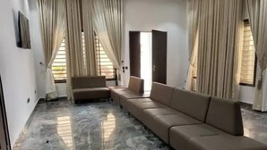 A spacious living room with a beige sectional sofa, a small sofa, and a coffee table. The room has a marble floor and large windows with beige curtains. - CITY DEN APARTMENT (Kaduna)