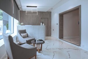 Uma área de recepção moderna, com um balcão branco, dois sofás cinzentos, piso de mármore e uma janela grande.