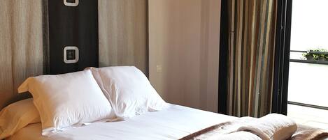 A hotel room with a large bed, a brown throw blanket, white pillows, and a window with curtains.