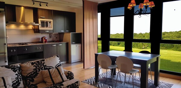 A modern kitchen with dark cabinetry, a microwave, and a refrigerator. There is a dining area with a table and chairs, and a view of a green landscape through large windows. - Apartamento Dúplex Golf Rioja Alta (Cirueña)