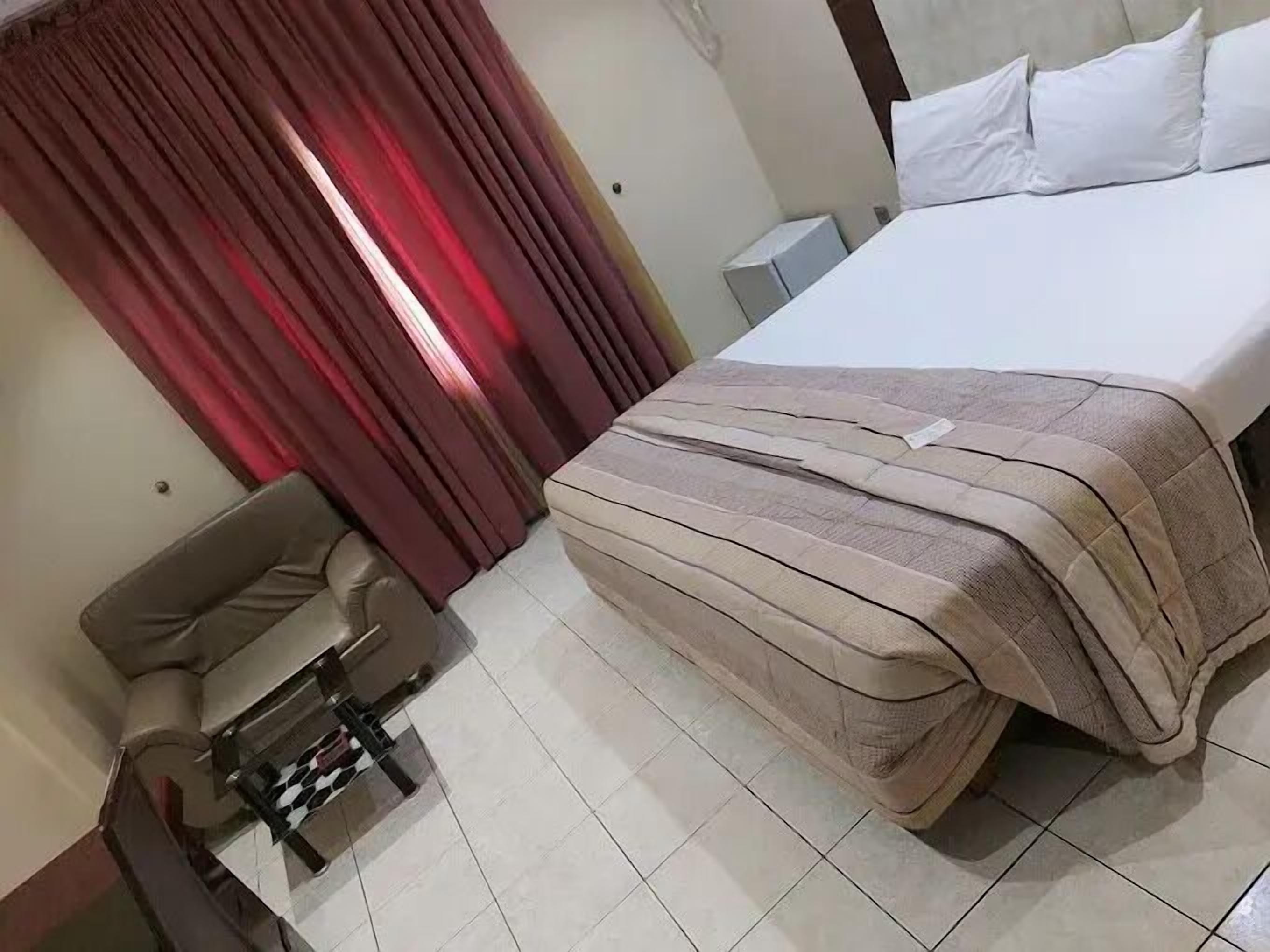 A hotel room with a bed, a chair, a desk, and a window with red curtains.