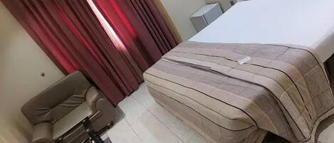 A hotel room with a bed, a chair, a desk, and a window with red curtains.
