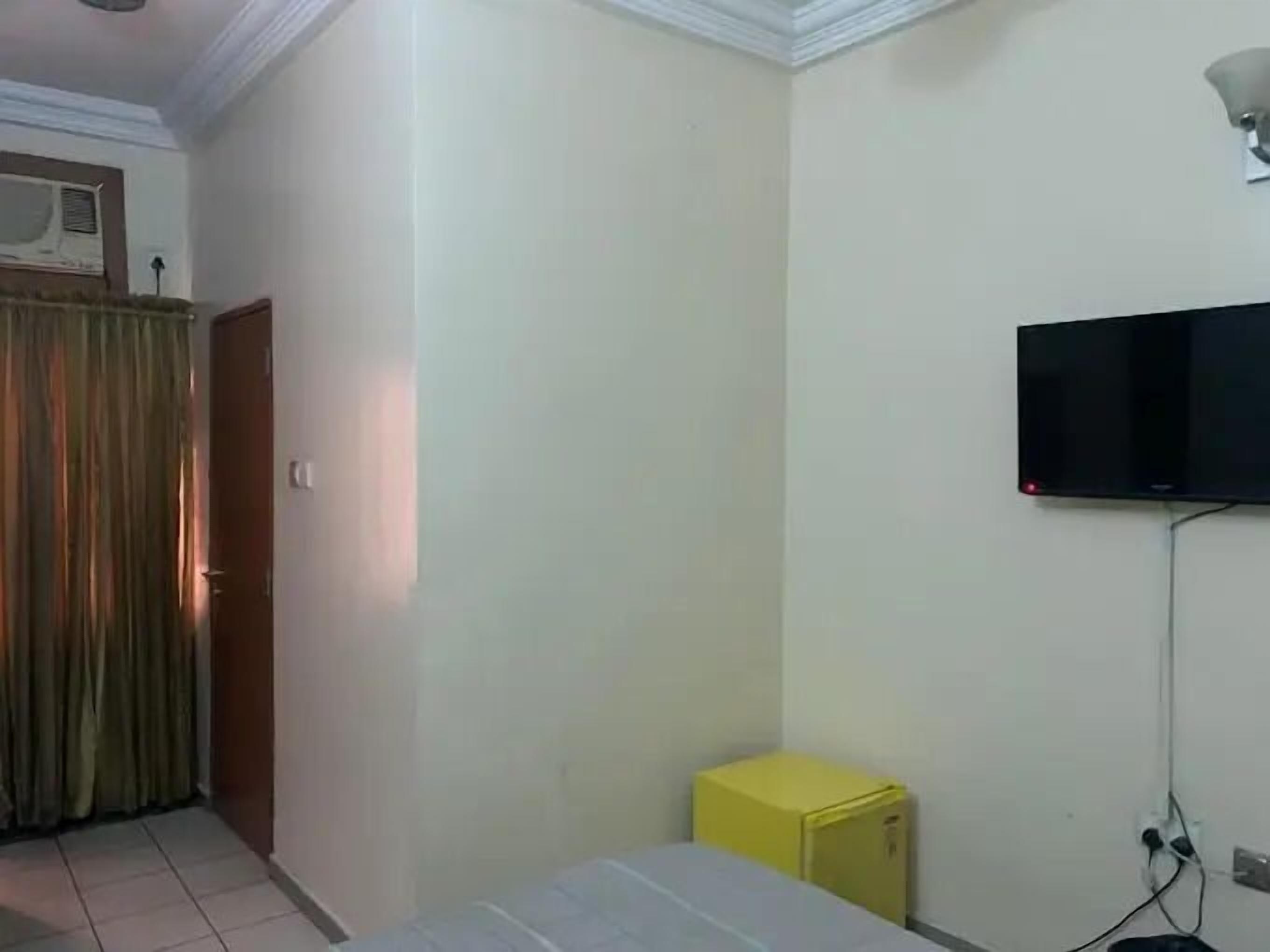 Une petite pièce comprenant un lit, un téléviseur fixé au mur, une petite armoire jaune et une porte avec des rideaux.