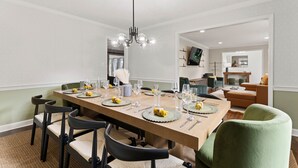 Un coin repas avec une longue table dressée pour six personnes, des chaises, un lustre et une vue sur un salon équipé d’un téléviseur.