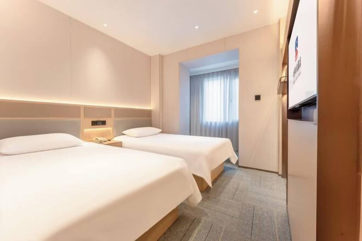 Ein Hotelzimmer mit zwei Betten, einem Nachttisch mit Lampe, einem Fenster mit Vorhängen und einem an der Wand befestigten Fernseher.
