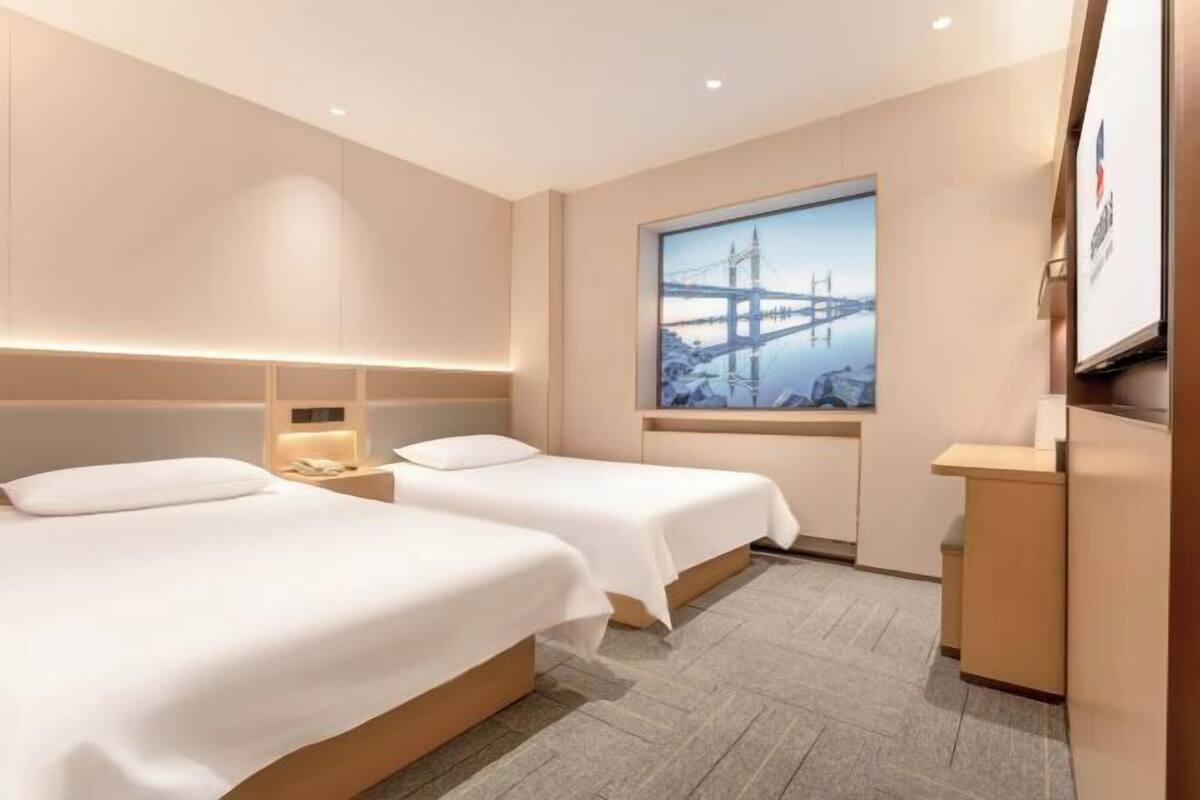 Ein Hotelzimmer mit zwei Betten, einem Schreibtisch, einem Fenster mit Blick auf die Stadt und einem Bild einer Brücke.