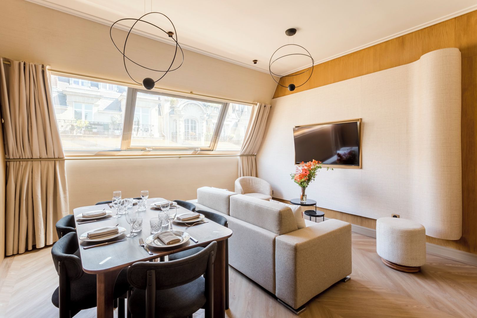 A modern hotel room with a dining area, a sofa set, a TV mounted on the wall, and a window with curtains.