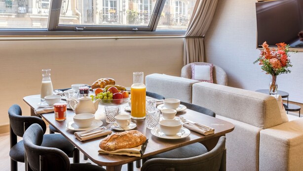 Une table de petit-déjeuner, des chaises et un canapé sont disposés dans une pièce dotée d’une grande fenêtre donnant sur un immeuble.