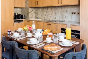 A breakfast setup with a table set for two, including plates, cups, a fruit bowl, a loaf of bread, and a bottle of orange juice.