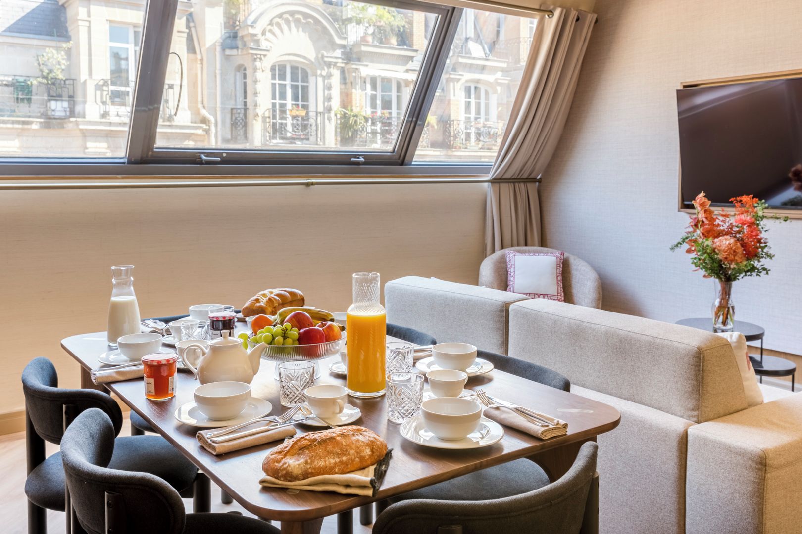 A breakfast setup with a table, chairs, and a sofa in a room with a large window overlooking a building.