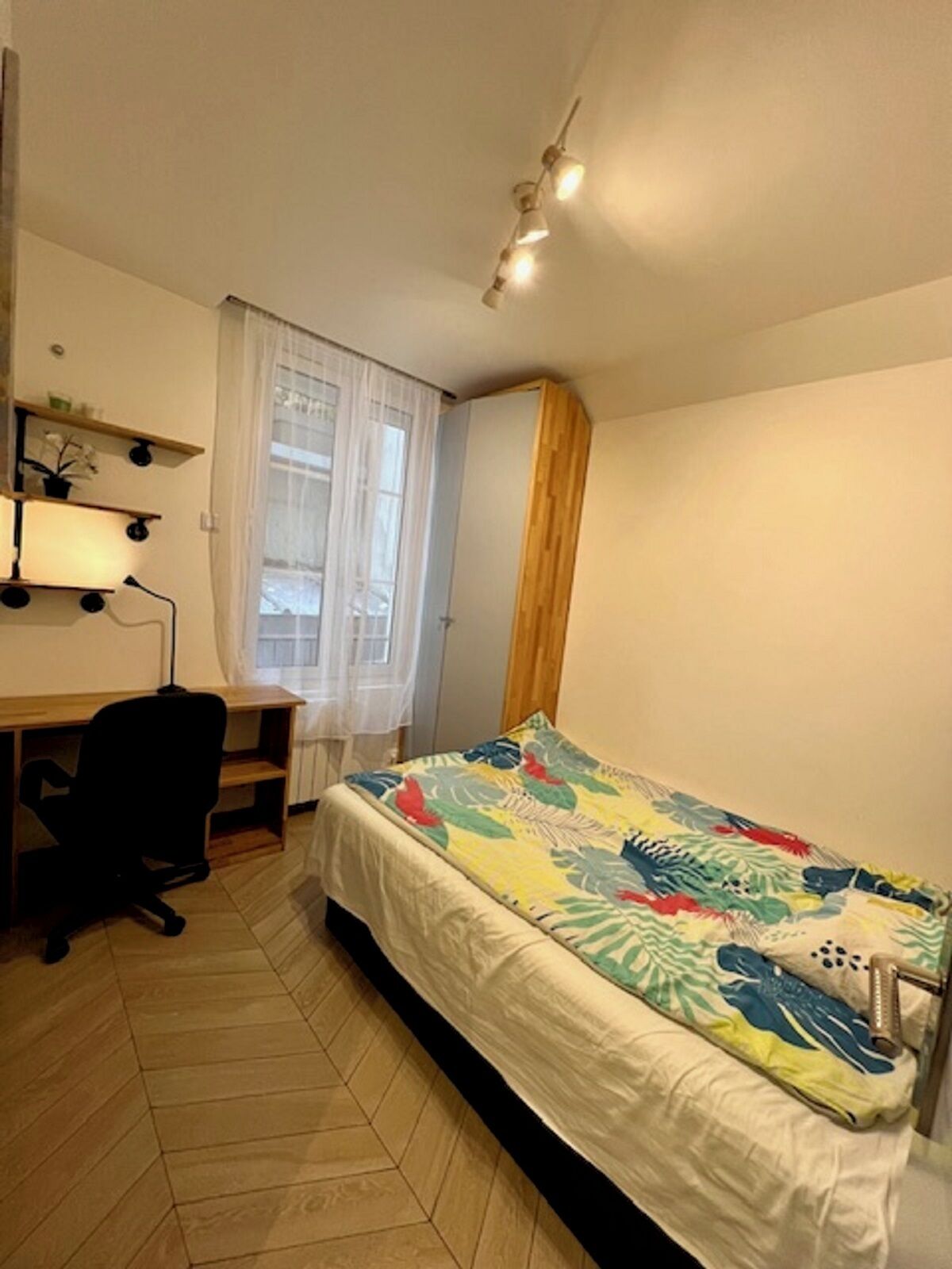 A small, well-lit bedroom with a bed, a desk, and a chair.