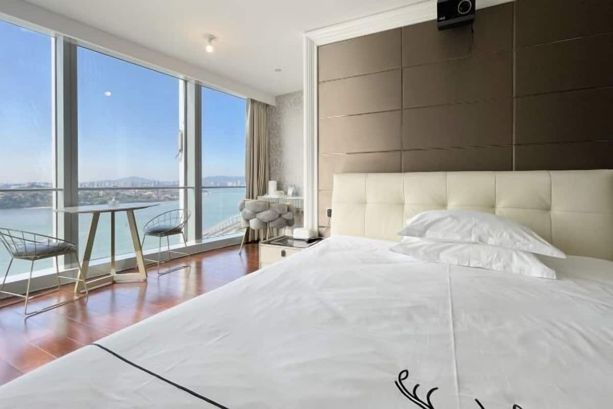 Un dormitorio con una cama grande, vistas a la ciudad, una mesita pequeña y una silla.