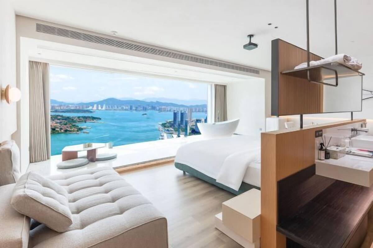 Una habitación de hotel moderna con una cama grande, un sofá, un escritorio y vistas a una ciudad costera.