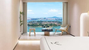 A modern hotel room with a large window offering a scenic view of a coastal town and mountains. - Xiamen IMAY Designer Seaview Apartment (Shimao Twin Towers Branch) (Xiamen)