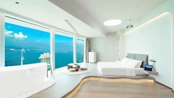 A modern hotel room with a large window, a bathtub, a bed, and a bedside table.
