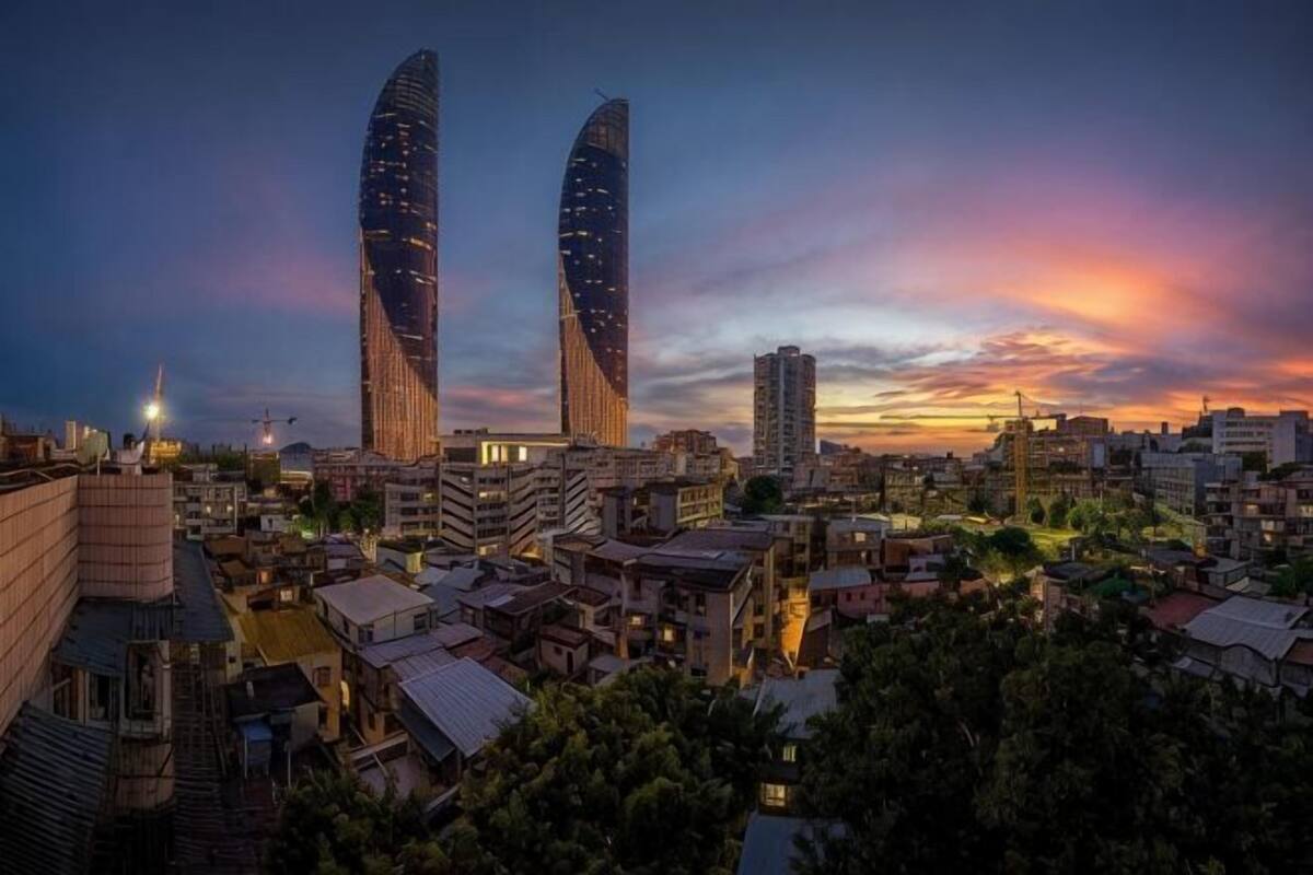 Un paisaje urbano con dos edificios destacados, una zona residencial densa y un cielo al atardecer.
