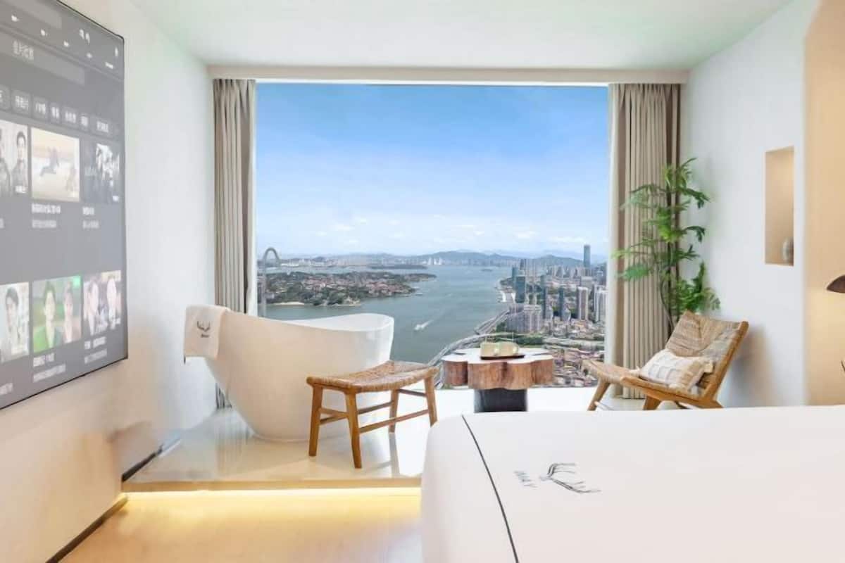 Una habitación de hotel moderna con una cama grande, una bañera blanca, una silla de madera, una mesita de madera pequeña y un ventanal con vistas a la ciudad.