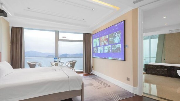 A hotel room with a bed, a large window, a wall-mounted TV, and a view of the sea. - Xiamen IMAY Designer Seaview Apartment (Shimao Twin Towers Branch) (Xiamen)