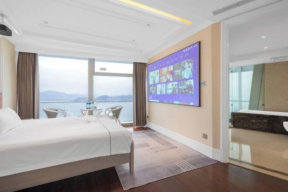 Habitación de hotel con cama, ventana grande, televisor montado en la pared y vista al mar.