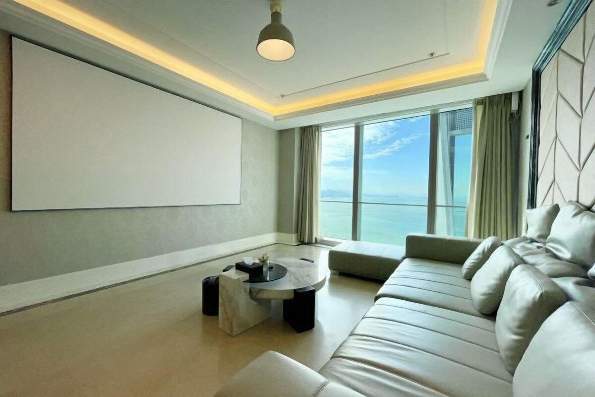 A modern living room with a large sofa, a coffee table, and a large window offering a view of the sea.