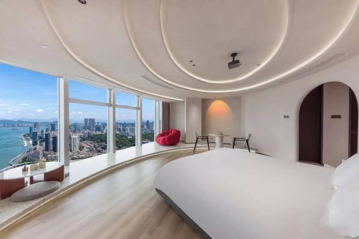 Una habitación de hotel moderna con una cama grande, un área de comedor, un sillón de peluche rojo y vistas a un paisaje urbano y una masa de agua.