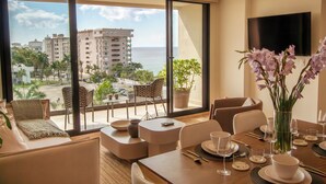 A hotel room with a large window offering a view of the ocean and buildings, a dining area with a table set for two, and a flat-screen TV mounted on the wall. - ATHIMAR Cozumel Condominium w/ Ocean View #406 (Cozumel)