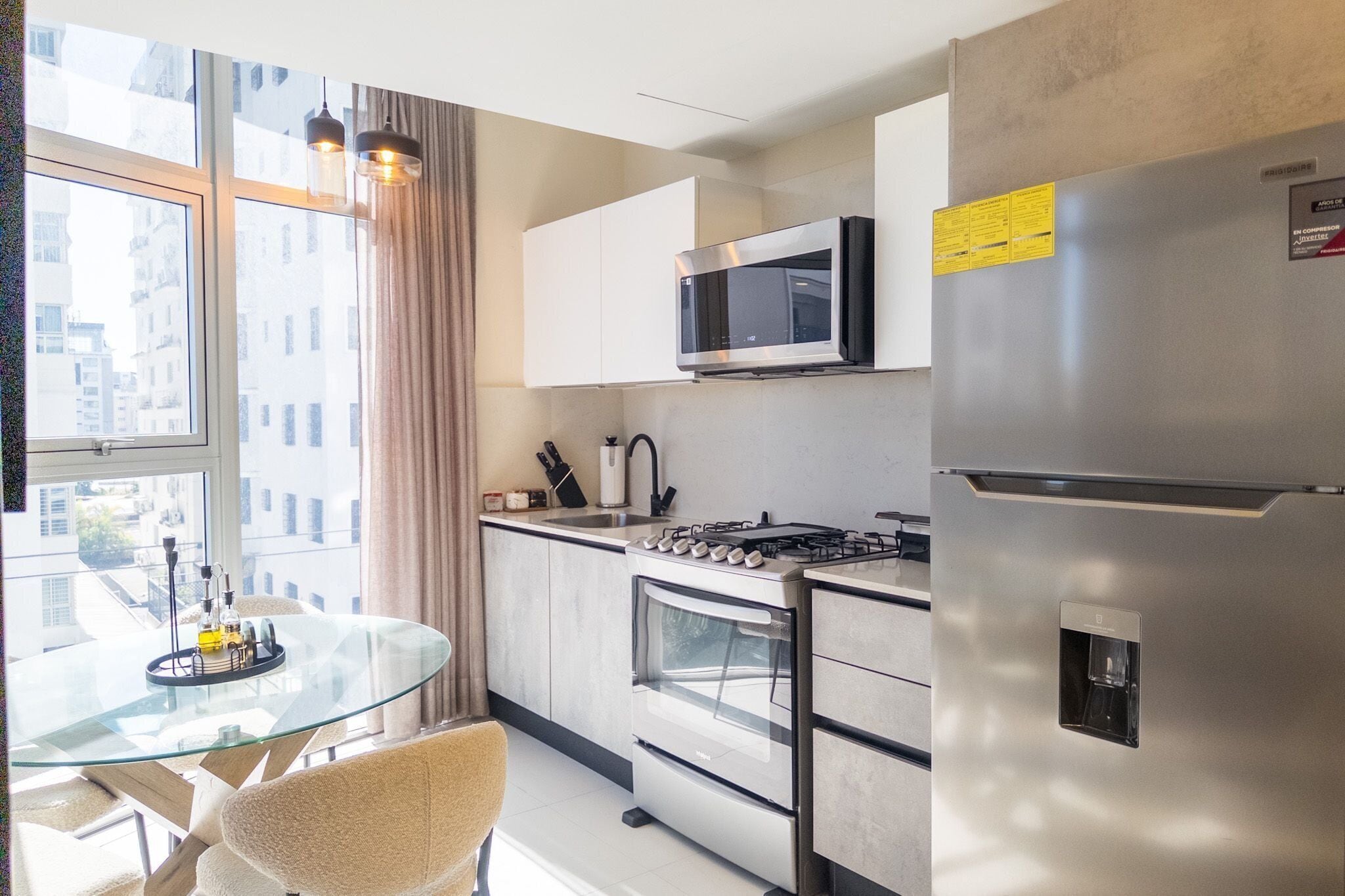 A modern kitchen with a glass dining table, stainless steel appliances, and a view of the city through large windows.