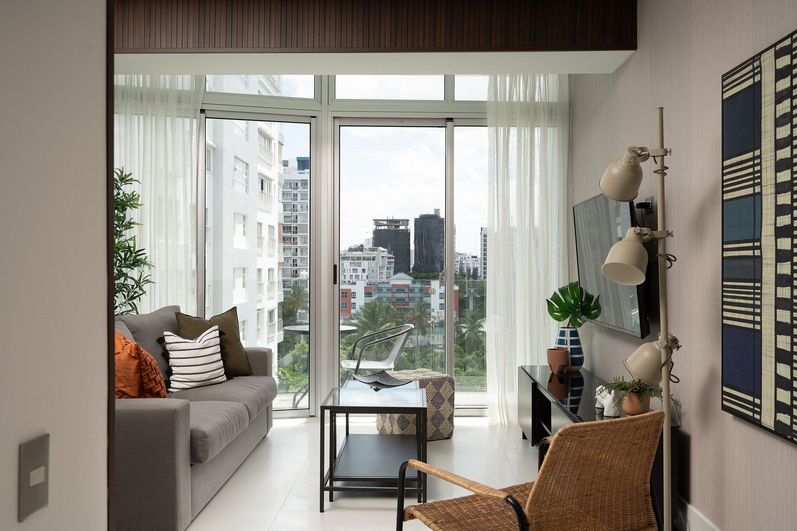 Ein modernes Wohnzimmer mit einem grauen Sofa, einem Glastisch und einem Korbstuhl. Ein großes Fenster bietet Blick auf die Stadt.