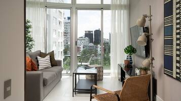 Ein modernes Wohnzimmer mit einem grauen Sofa, einem Glastisch und einem Korbstuhl. Ein großes Fenster bietet Blick auf die Stadt.