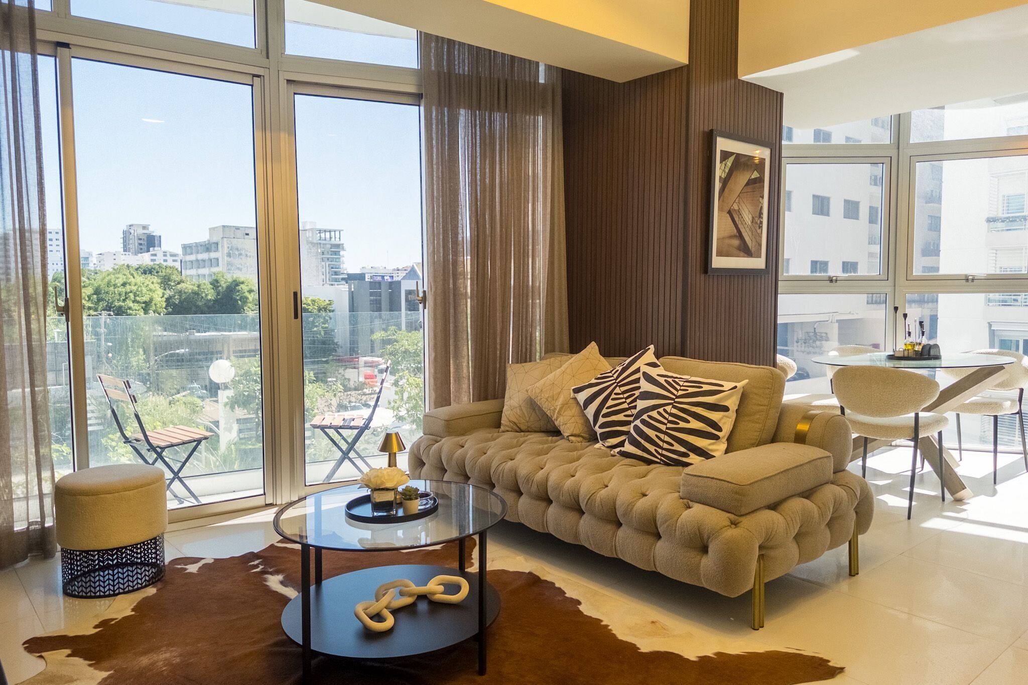 Una sala de estar moderna con sofá, reposapiés, mesa de centro y una mesa de vidrio. Amplios ventanales ofrecen vistas a la ciudad.