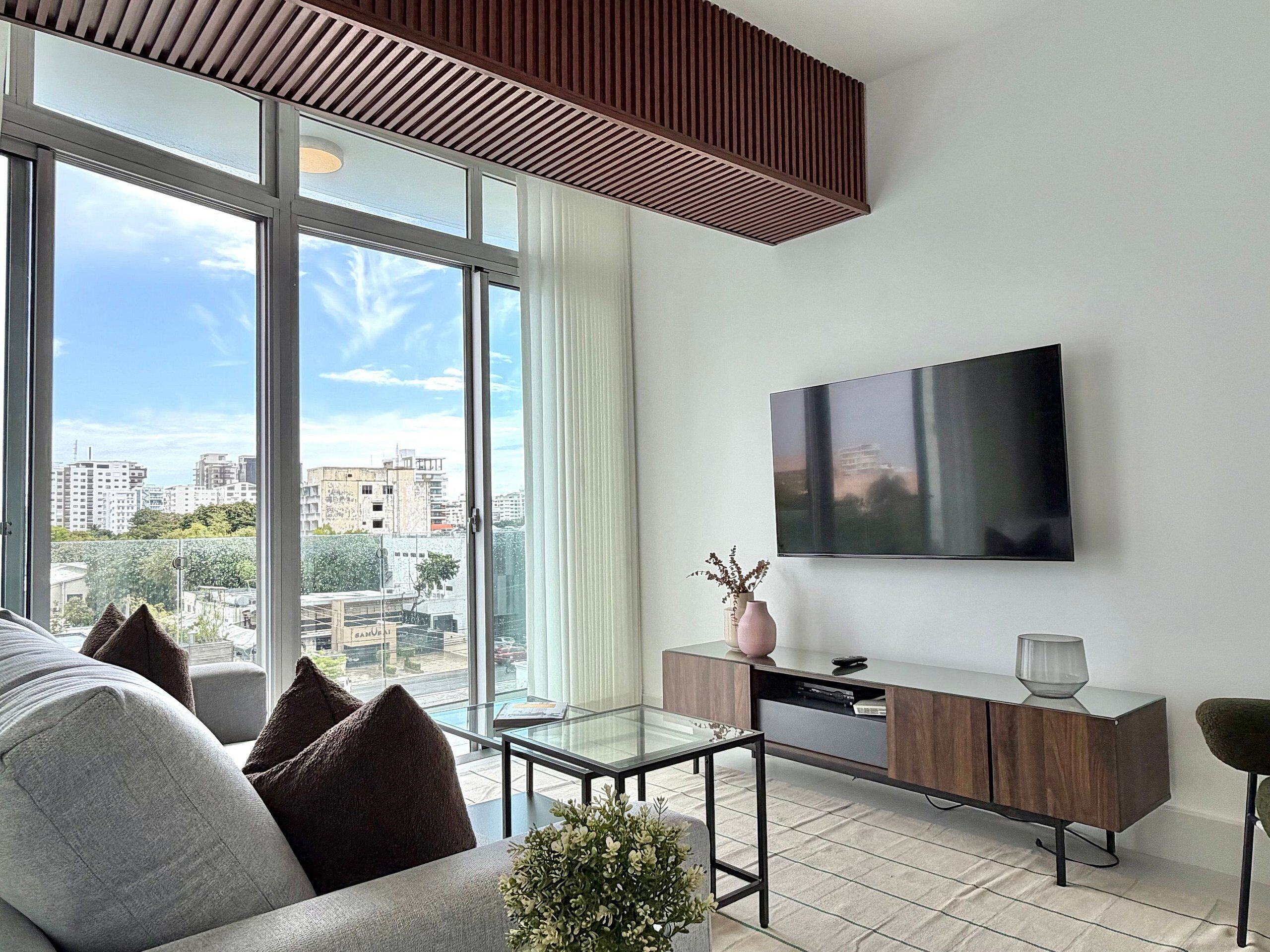 A modern living room with a large window, a flat-screen TV, a glass coffee table, and a minimalist console table.