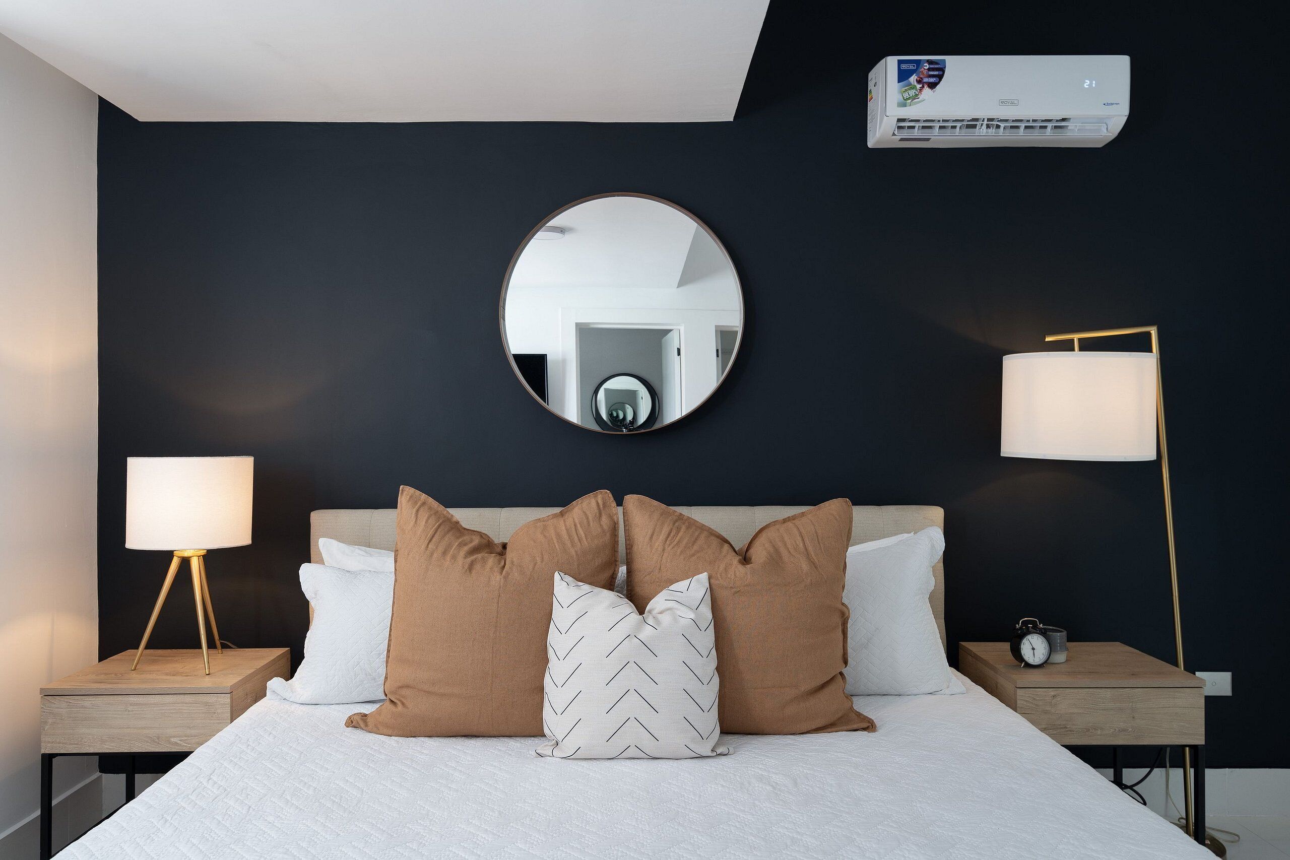 A neatly made bed with a white and brown pillow arrangement, a round mirror above, and two bedside tables with lamps and an alarm clock.