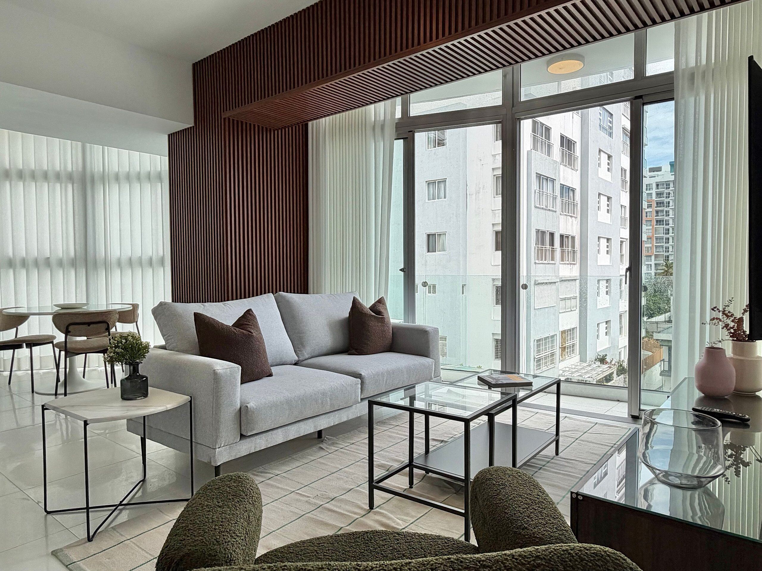 A modern living room with a sofa, coffee table, and chairs, large windows with a city view, and a wooden accent wall.
