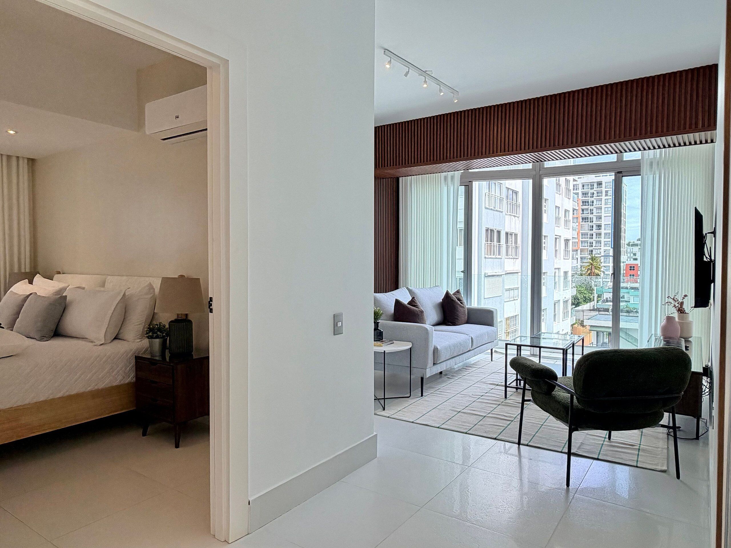 Un apartamento moderno con cama, sofá, zona de comedor y grandes ventanales con vistas a la ciudad.