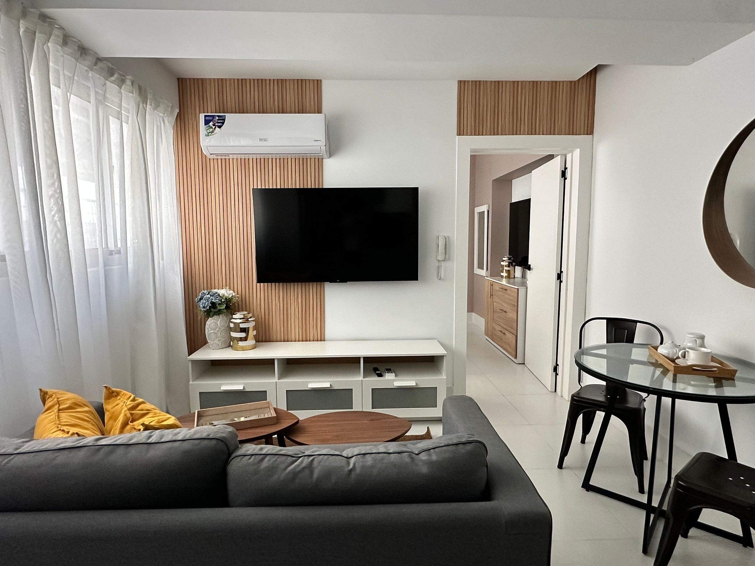A modern living room with a grey sofa, a flat-screen TV, a wooden accent wall, a dining area with a glass table and black chairs, and a kitchen in the background.