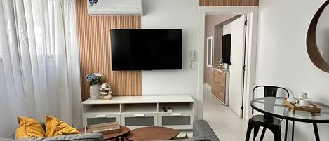 A modern living room with a grey sofa, a flat-screen TV, a wooden accent wall, a dining area with a glass table and black chairs, and a kitchen in the background.