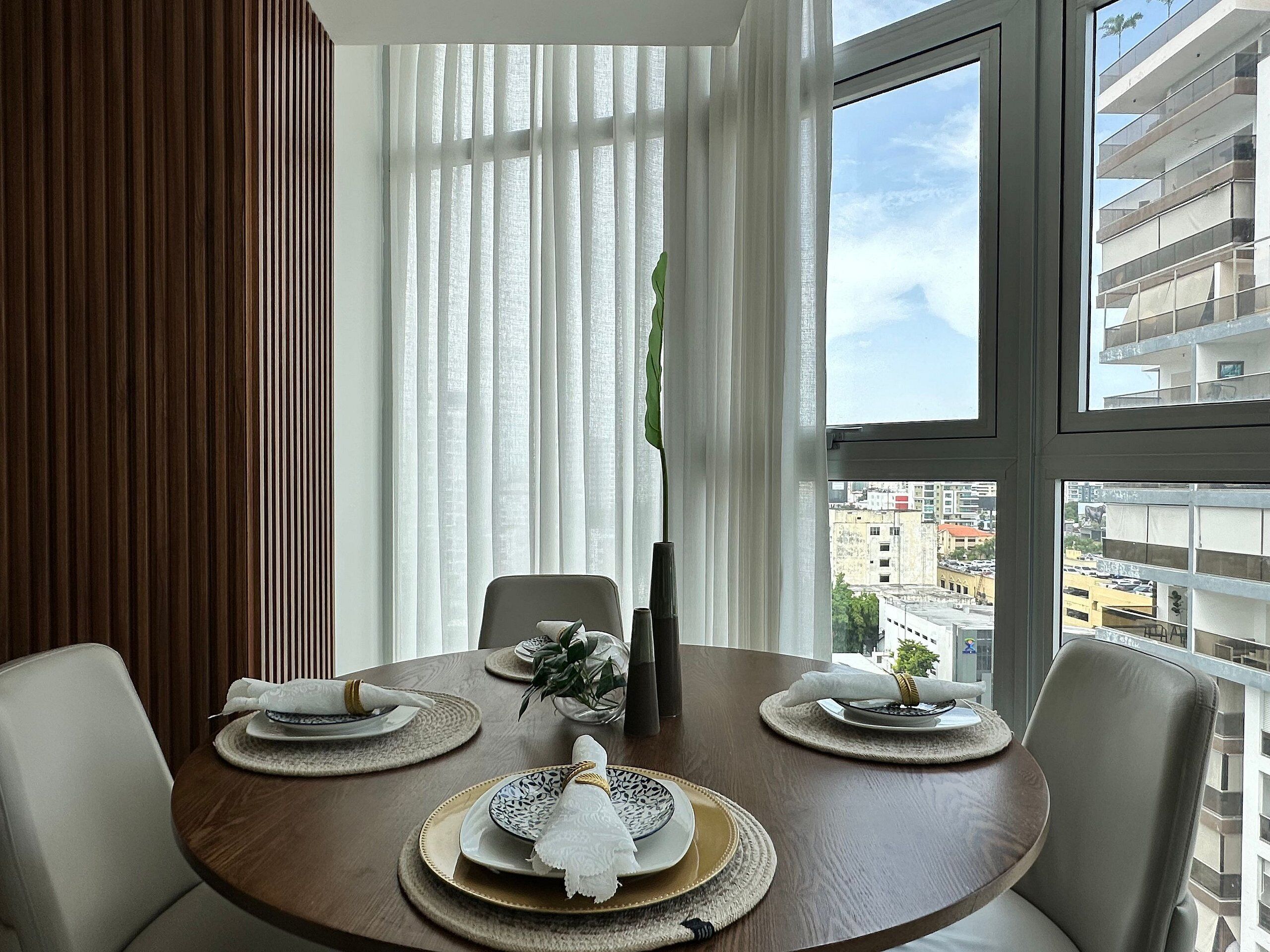 Ein Essbereich mit einem runden Tisch, gedeckt für vier Personen, einem Fenster mit Jalousien und Blick auf die Stadtlandschaft.