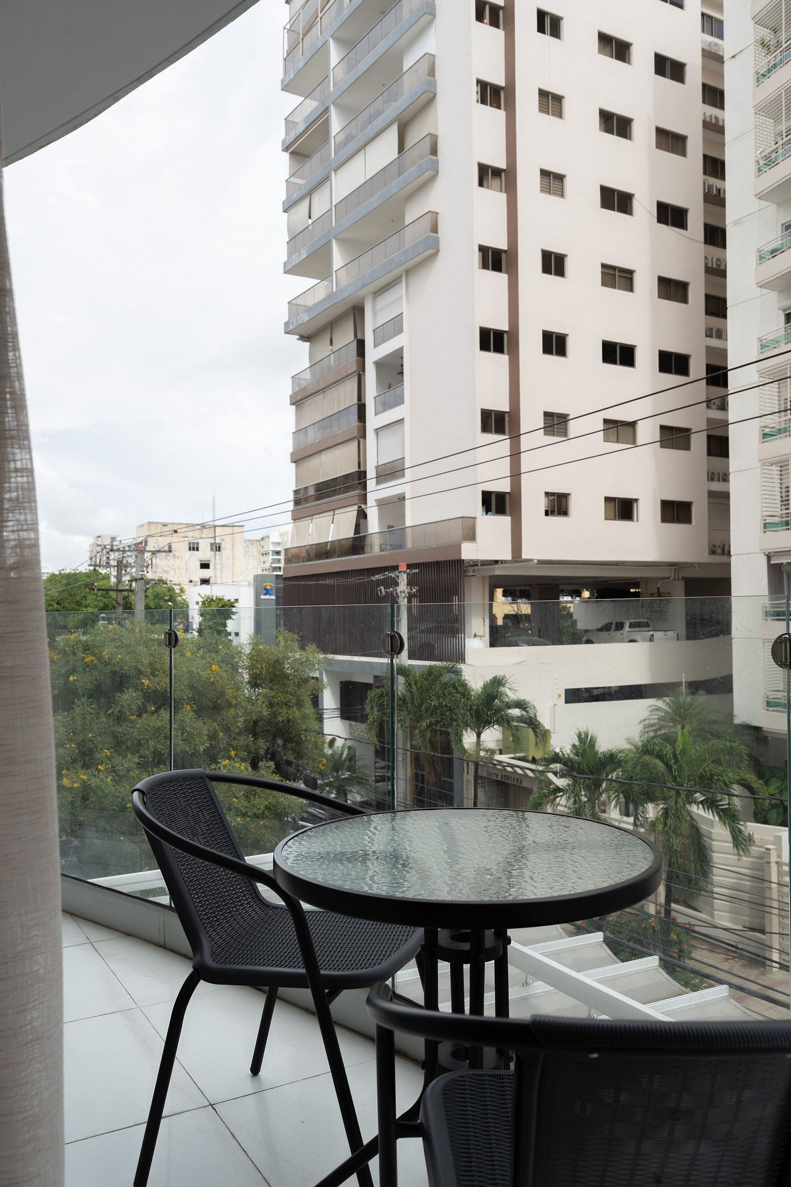 Un balcón con una mesa y sillas de vidrio, con vistas a un paisaje urbano con edificios altos y zonas verdes.