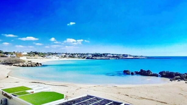 Une plage à l’eau d’un bleu limpide, avec un rivage sablonneux et une ville qui se devine au loin.
