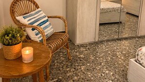 Une pièce avec un fauteuil en osier, une petite table sur laquelle est posée une bougie, et un miroir qui reflète un lit et une table de chevet.