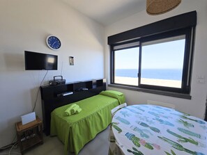 A bedroom with a bed, a TV, a desk, and a window with a beach view. - Dammuso - Campobello - RENT'CA (Pantelleria)