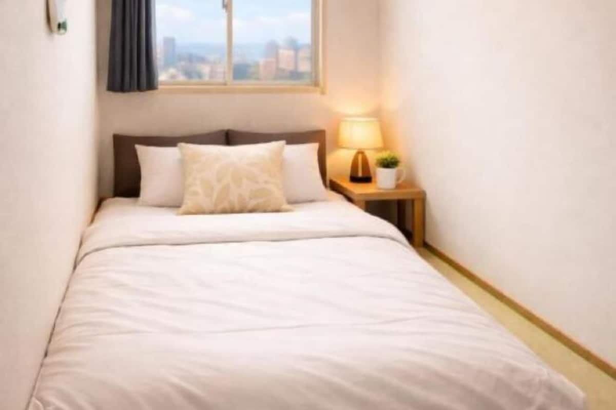 A hotel room with a bed, a nightstand with a lamp, and a window with a view of buildings.