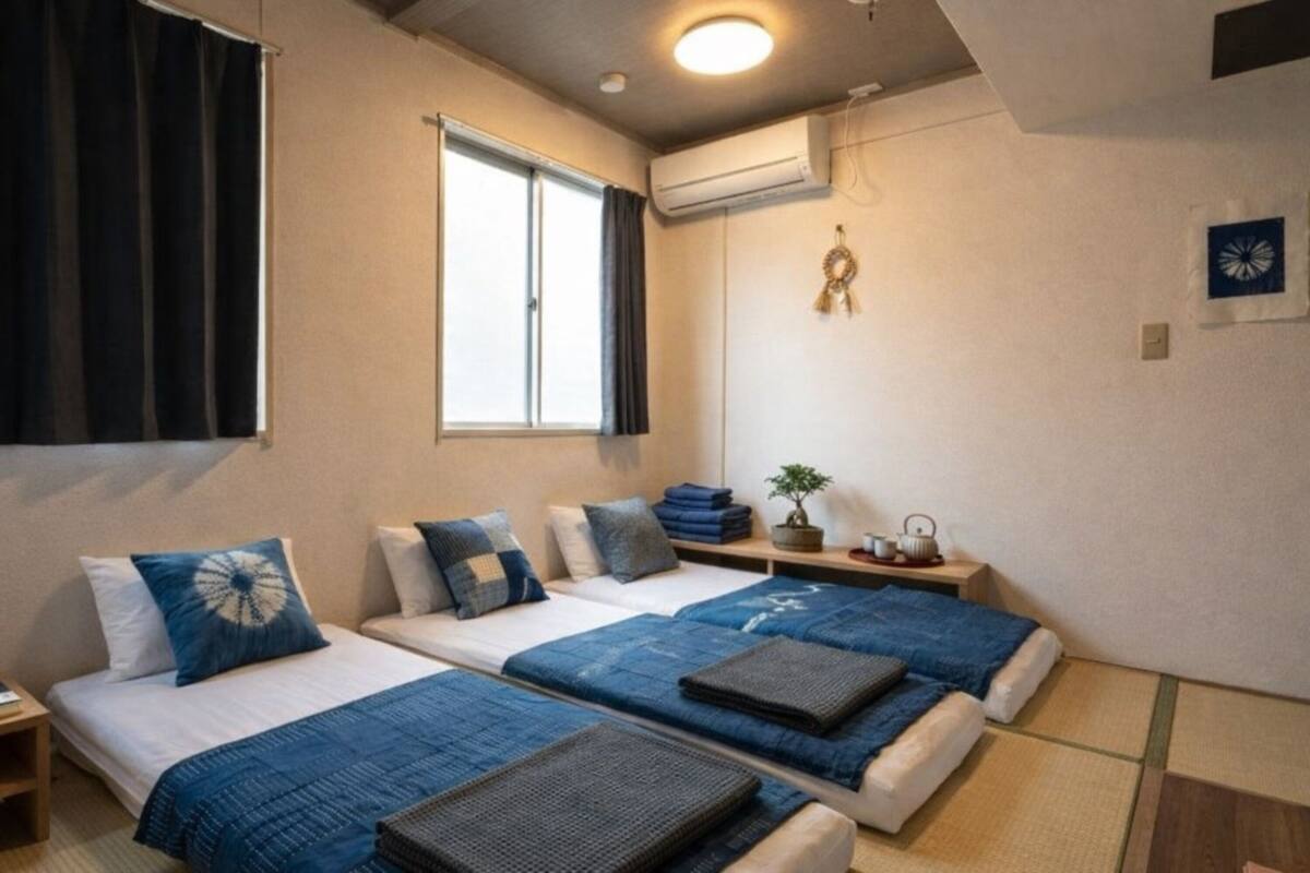 A hotel room with two beds, a window, an air conditioner, a small table with a plant, and a wall decoration.